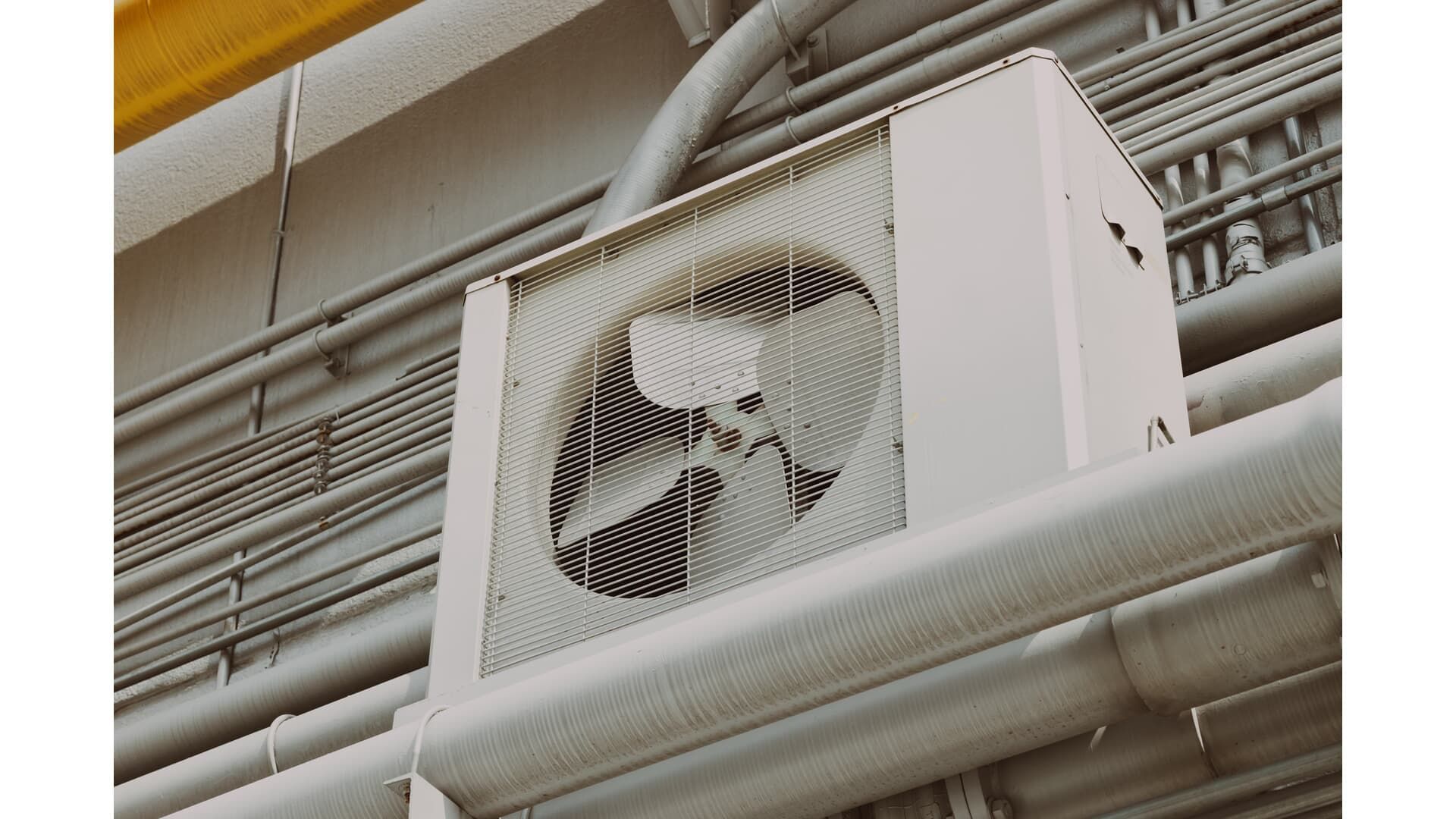 A white outdoor air conditioning unit mounted on a wall with pipes running alongside it.