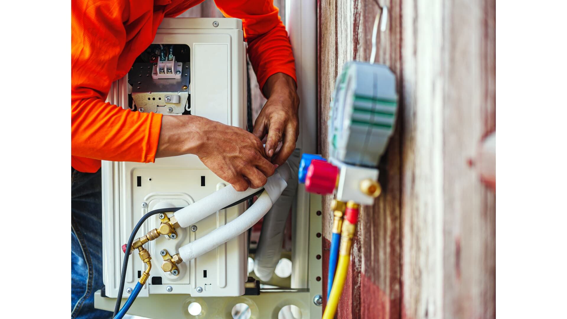 A technician in an orange shirt works on an outdoor air conditioning unit connected to a refrigerant pressure gauge.
