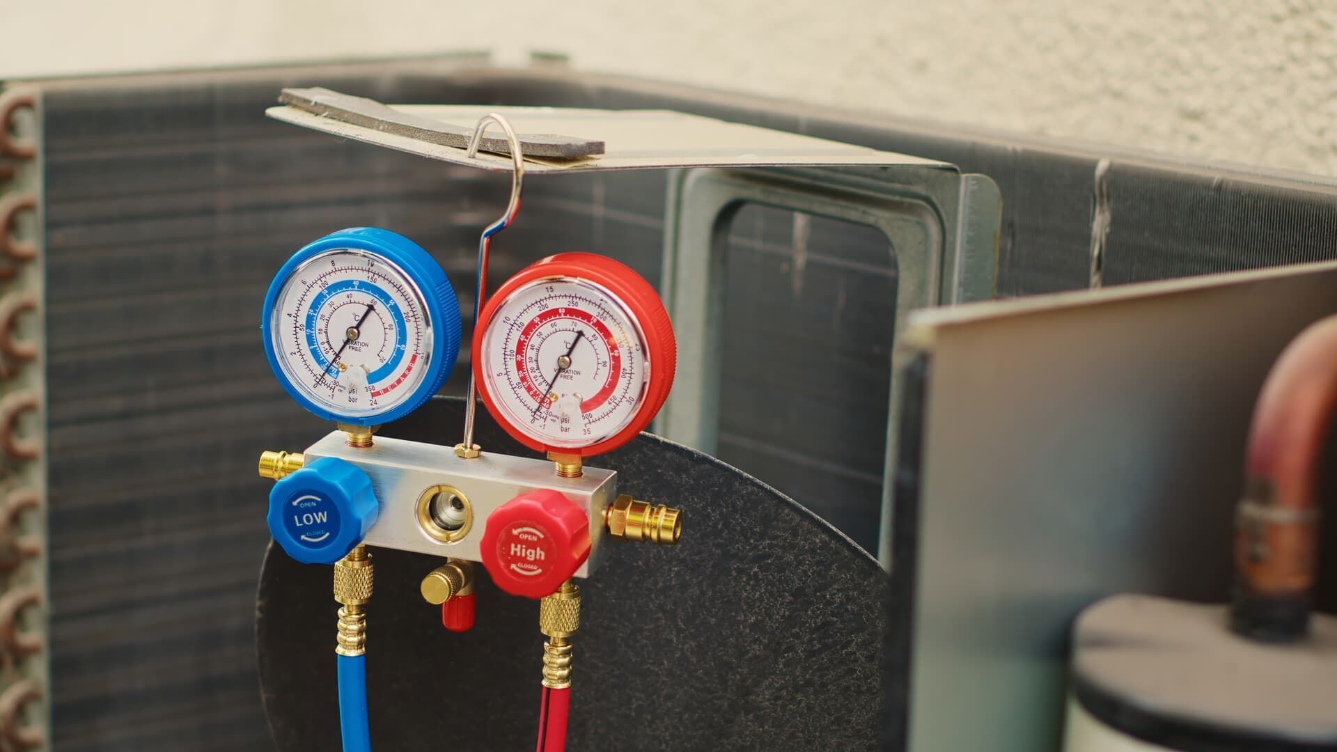 Manifold gauges with red and blue dials connected to an HVAC unit's copper piping for maintenance.