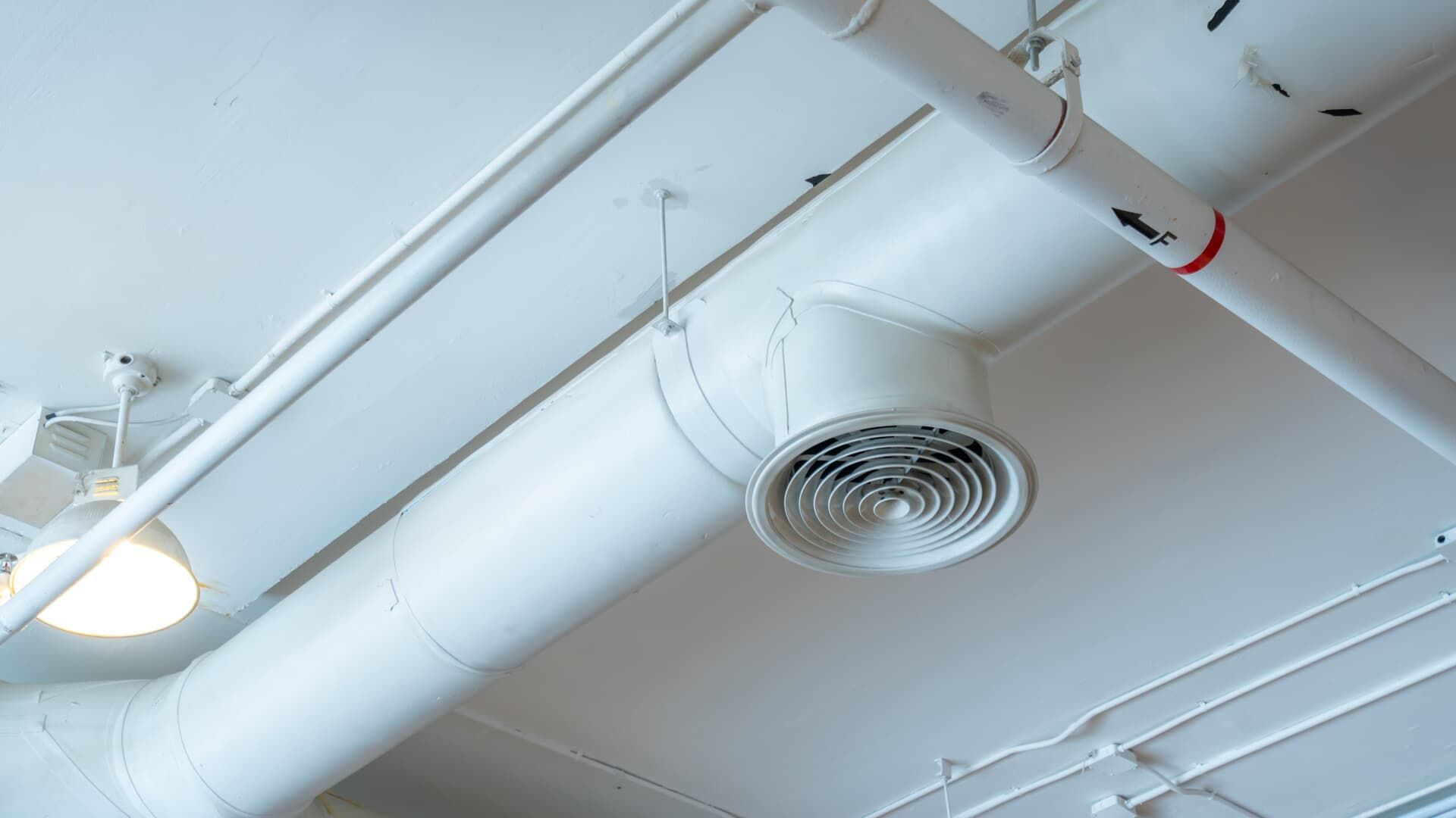A white industrial ventilation duct and a circular ceiling air diffuser suspended from a white ceiling with light fixtures.