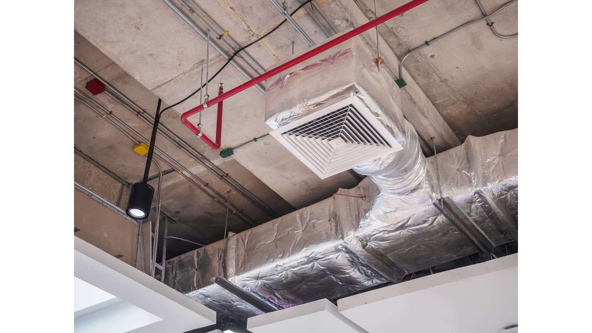 Exposed concrete ceiling with a silver ventilation duct, a square air diffuser, and a red fire suppression pipe.