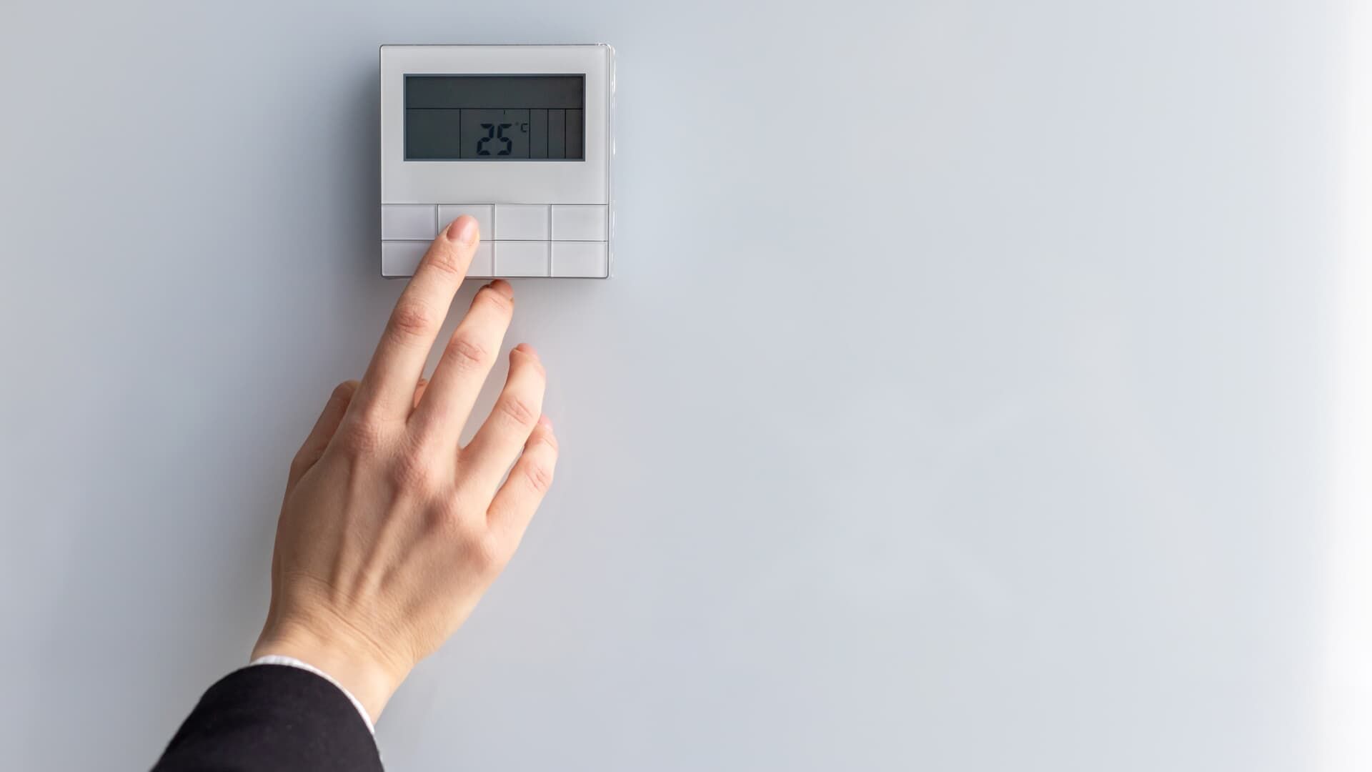 A hand in a dark sleeve presses a button on a wall-mounted thermostat displaying 25 degrees against a white background.