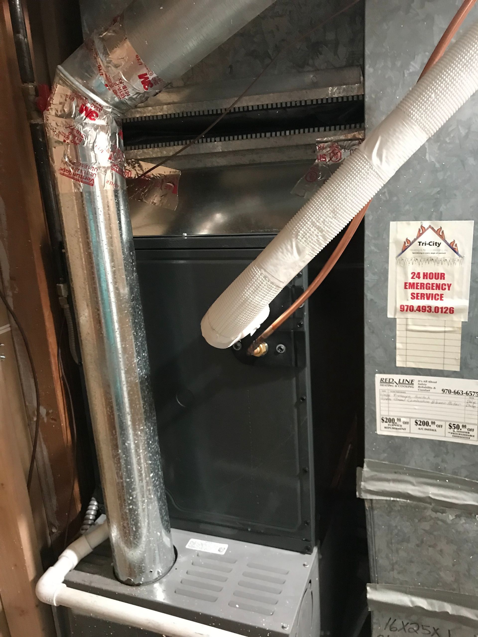 A basement furnace system with metal ductwork, white PVC piping, and an HVAC service sticker on the side unit.