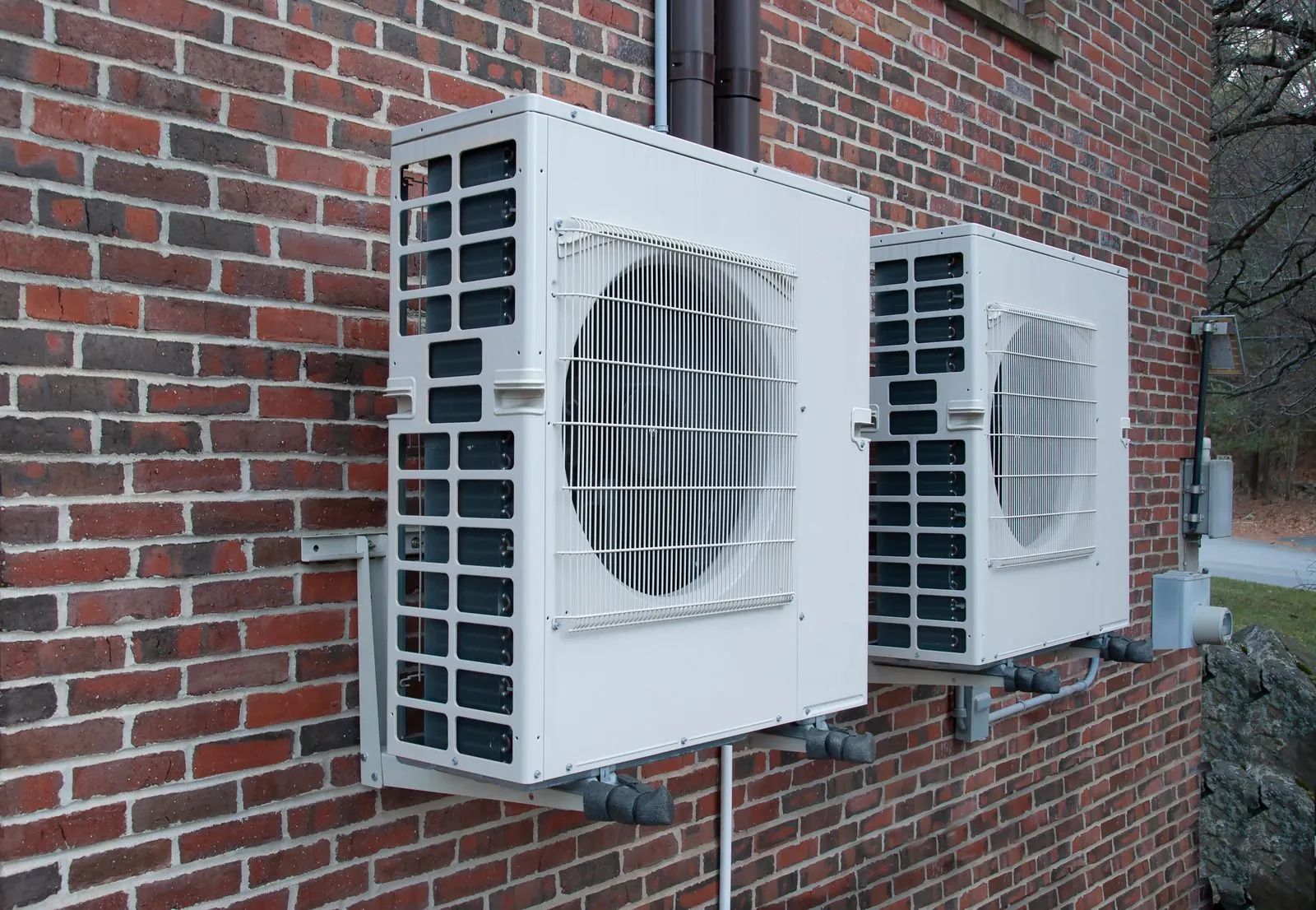 Two white outdoor air conditioning units mounted on a red brick wall.