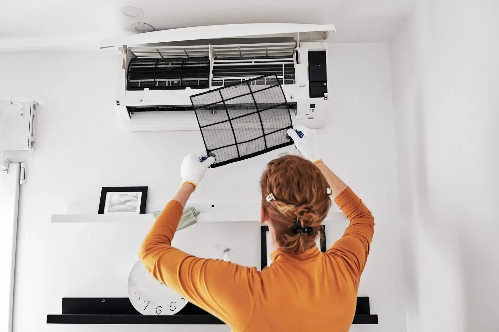 A person wearing white gloves removes a dust filter from a wall-mounted air conditioner in a bright, modern room.