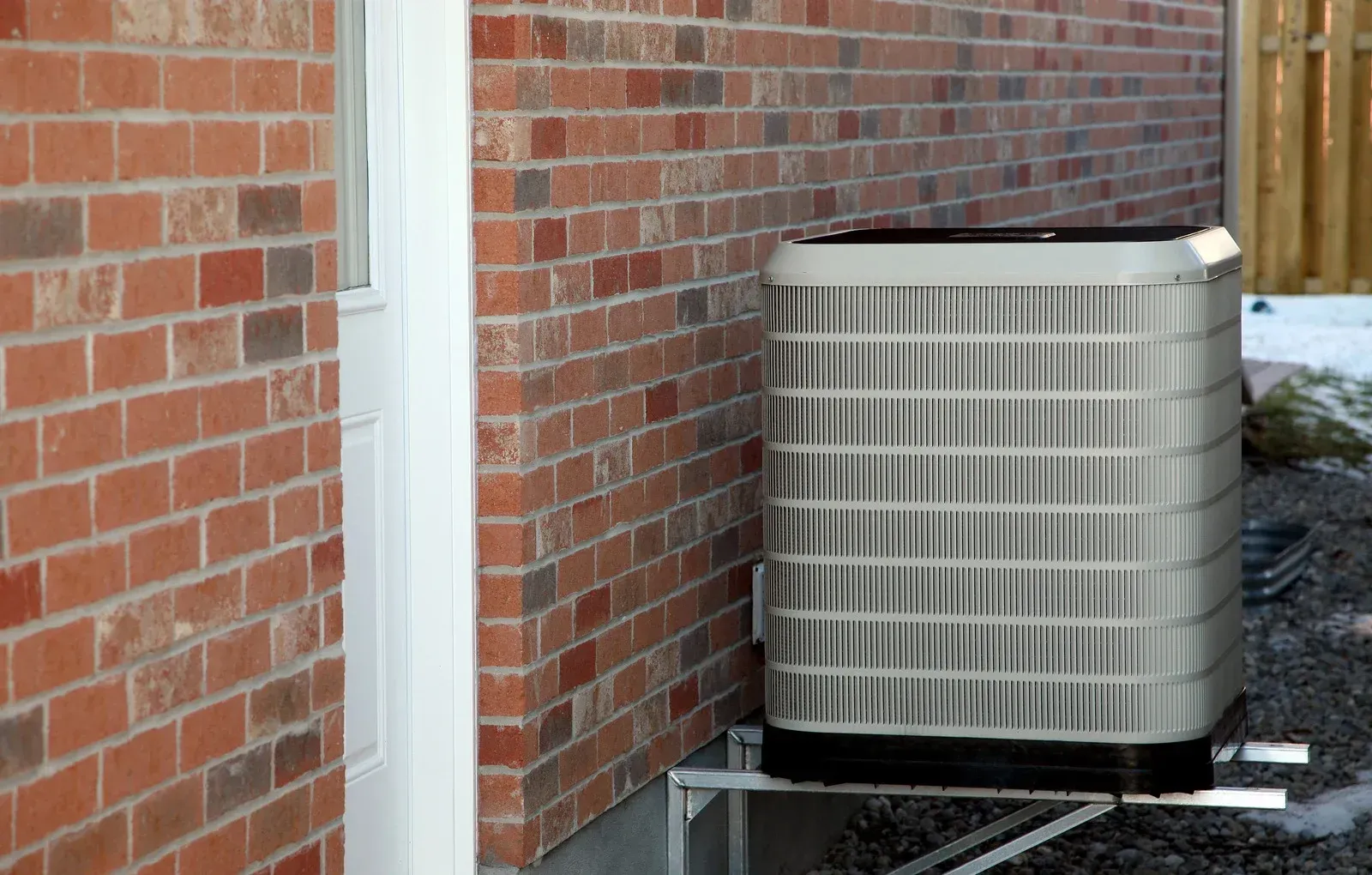 An outdoor HVAC condenser unit sits on a metal stand beside a red brick wall near a doorway.
