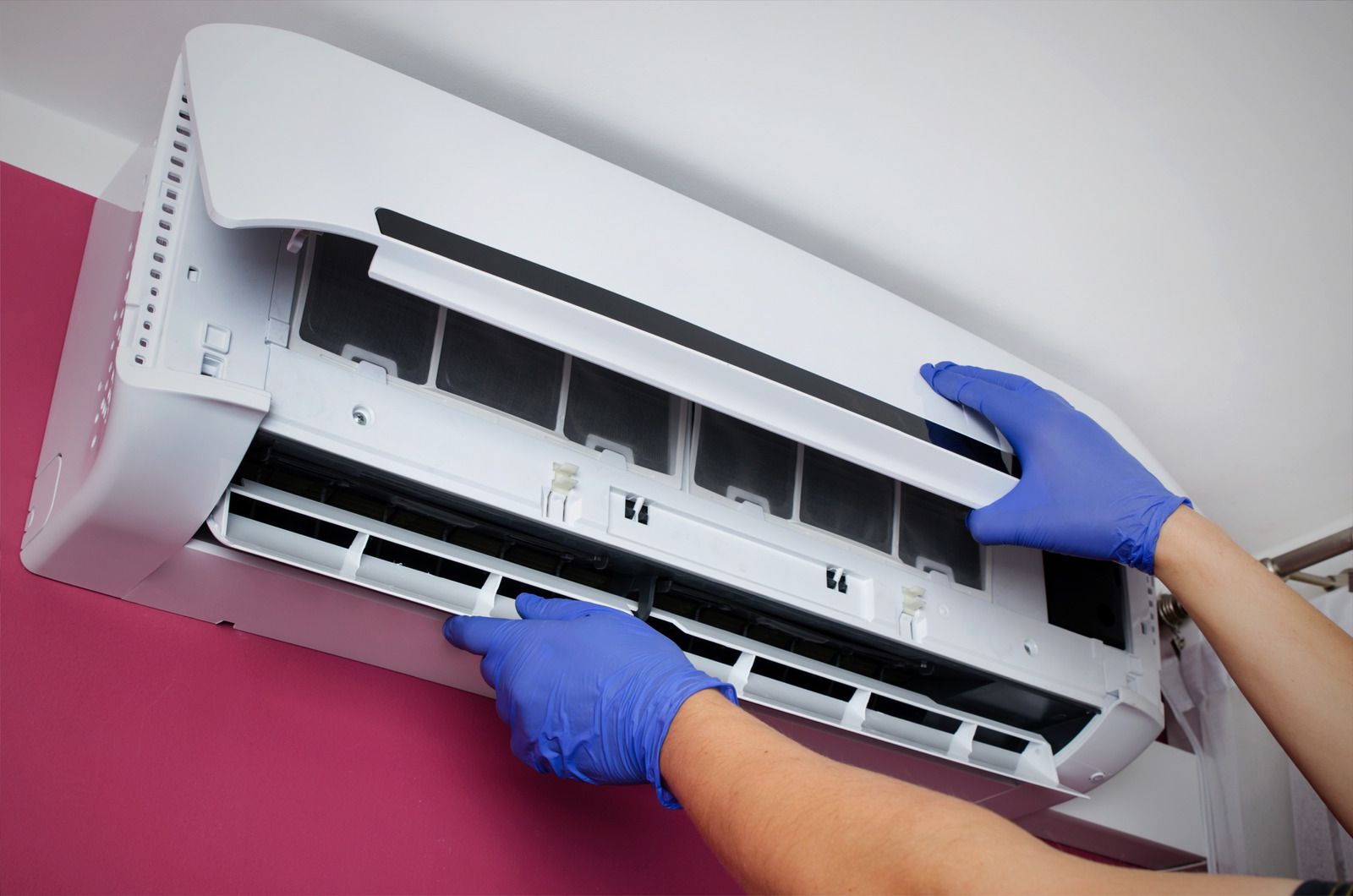 A technician wearing blue gloves opens the front panel of a wall-mounted air conditioner to perform maintenance.