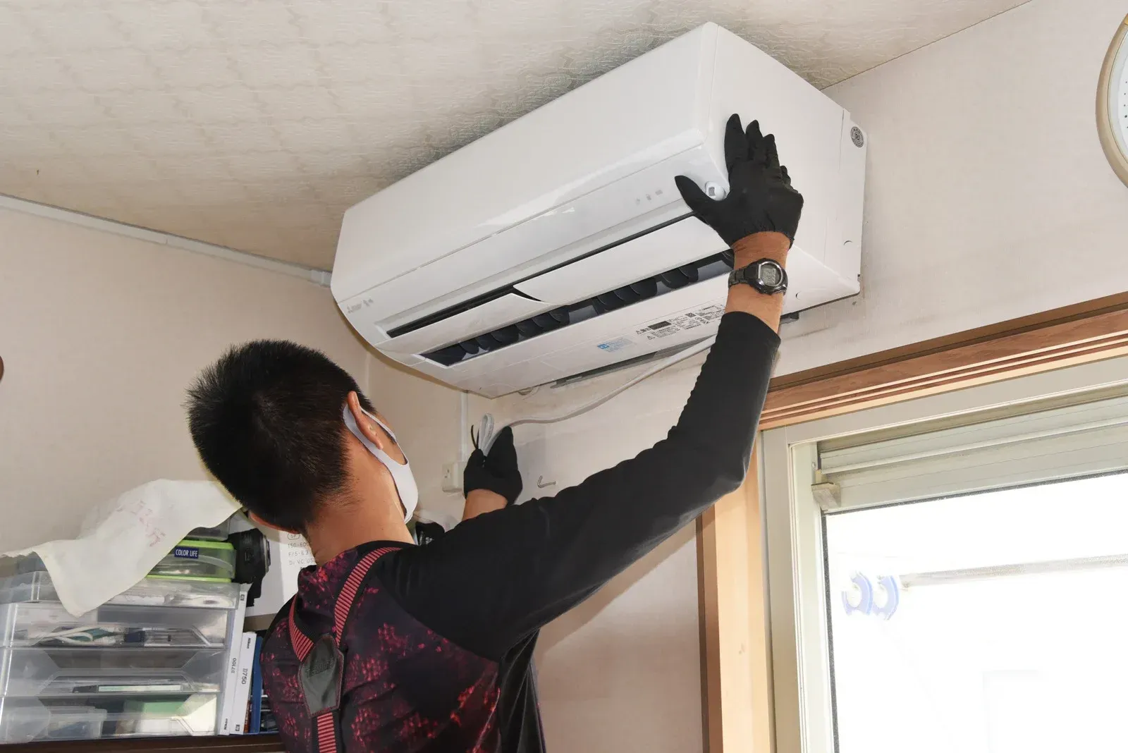 A technician in black work clothes installing a white air conditioning unit on a room wall.