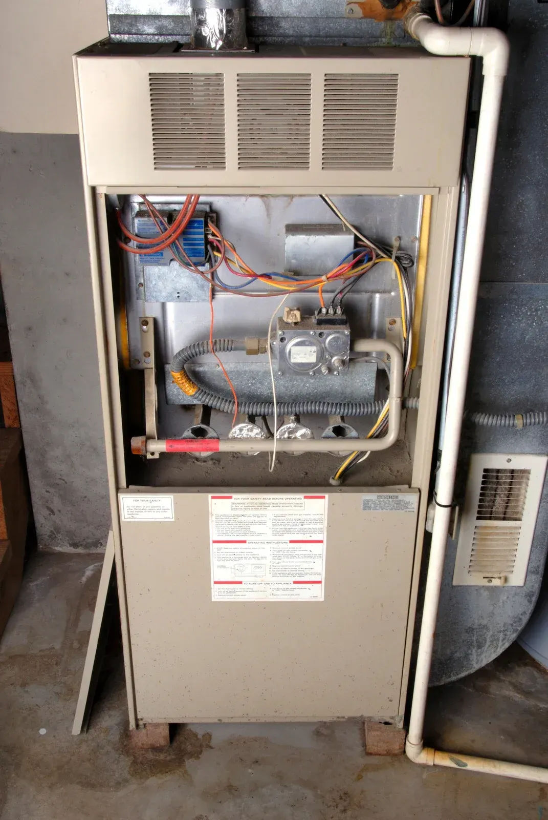 Open beige gas furnace in a basement showing interior burner components, wiring, and a white PVC drain pipe.