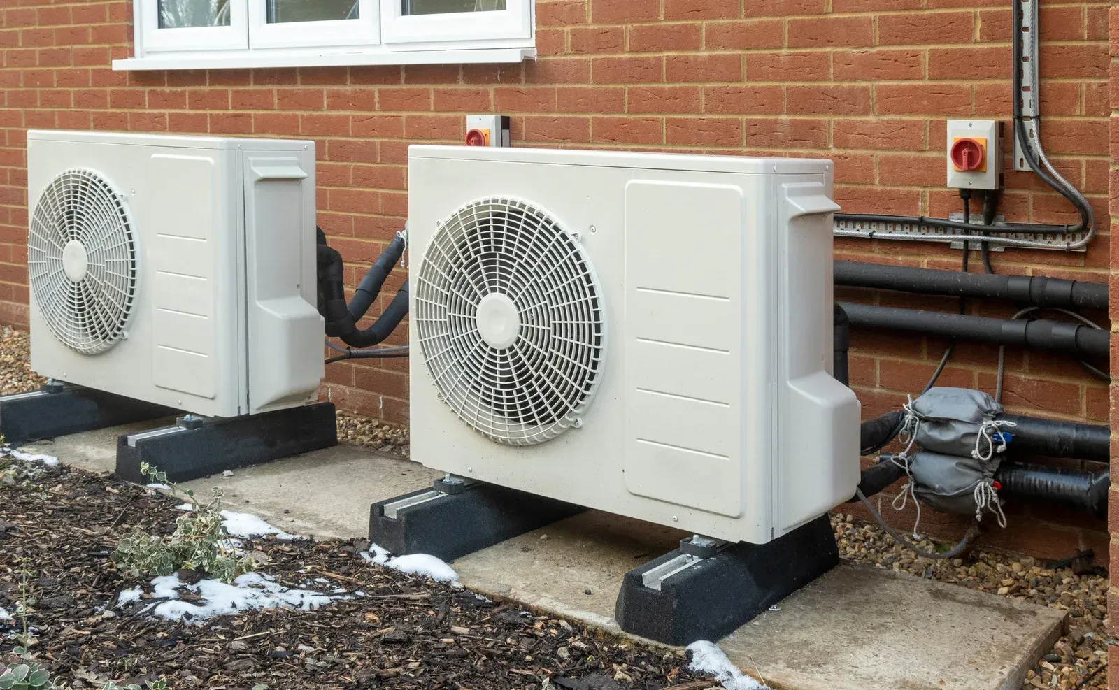 Two light-colored heat pump units installed on concrete blocks against a brick wall outside a building.