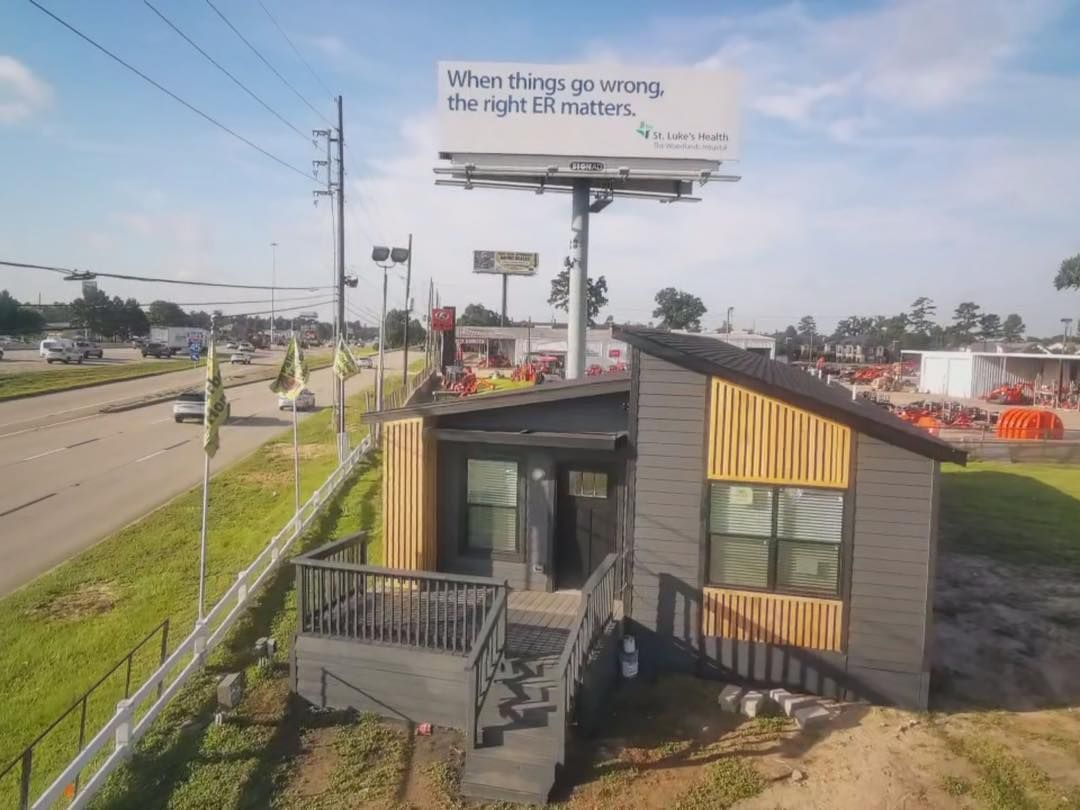 Small house next to road, with billboard overhead. House is gray with wooden accent.