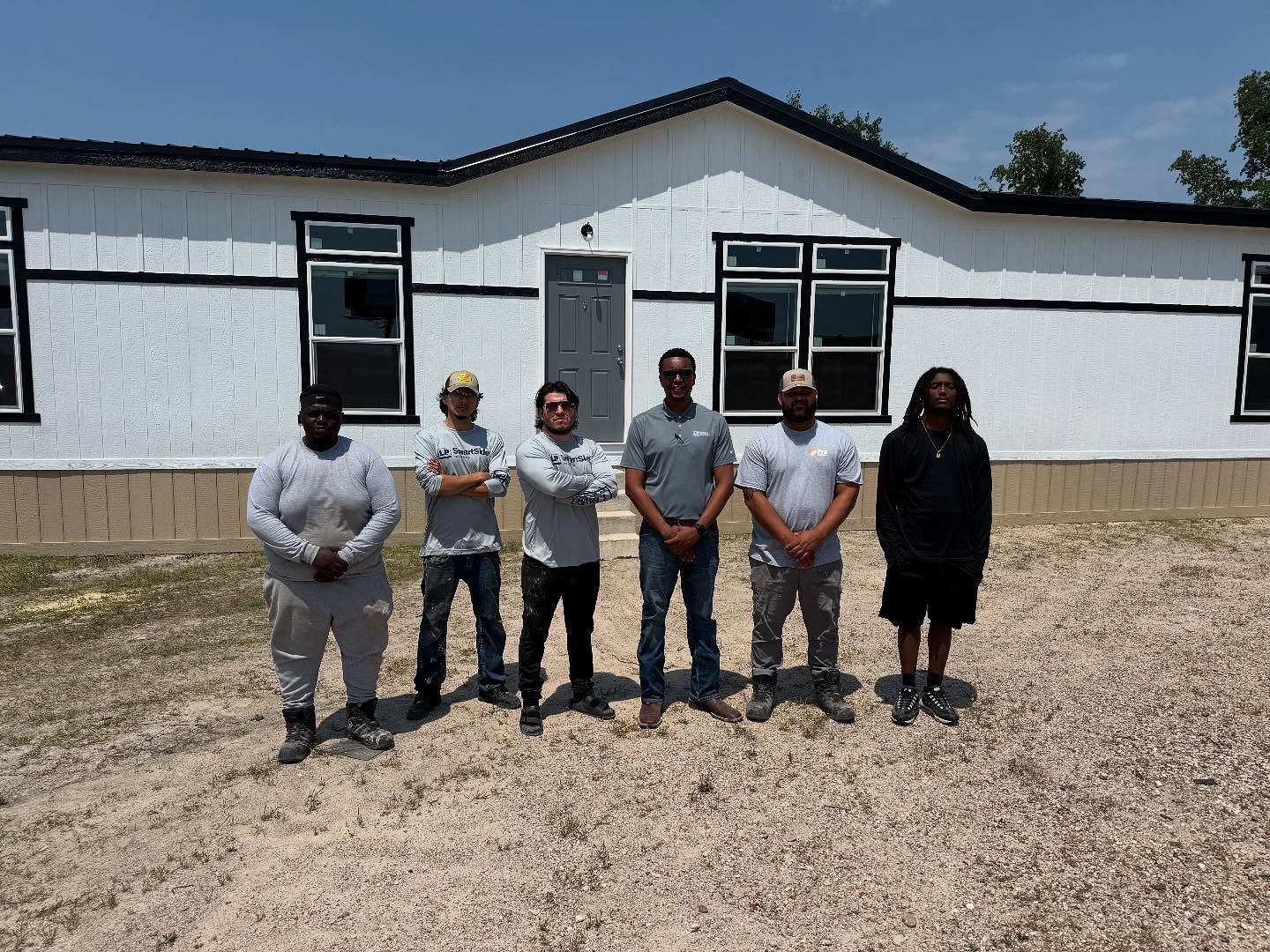 Group of men in front of a white house with black trim, standing on a gravel surface.