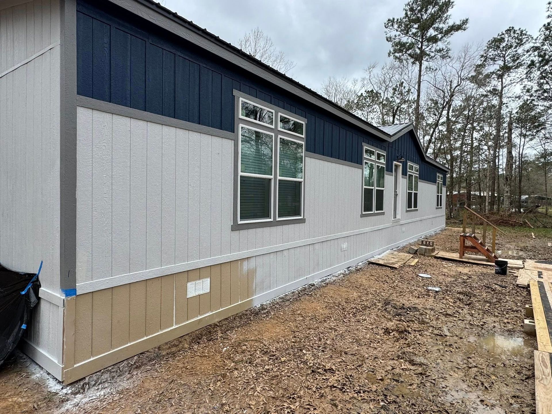Exterior view of a house with gray siding, blue trim, and windows. Construction materials on the ground.