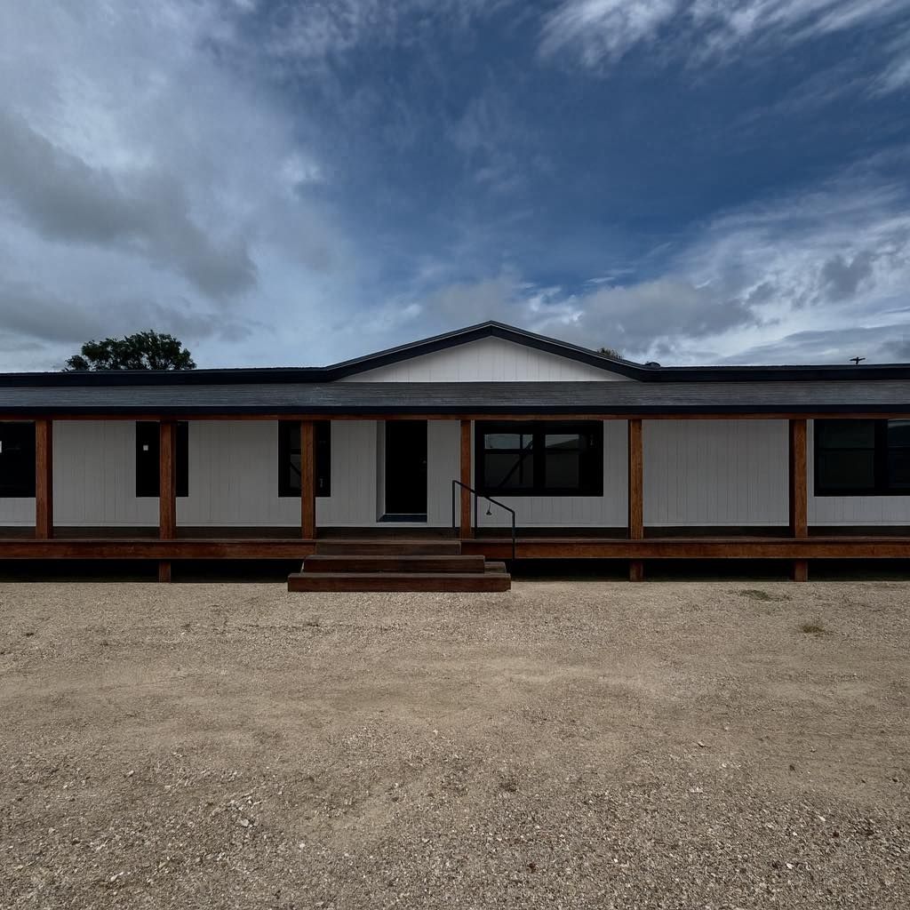 White house with a dark roof and porch, set on a gravel lot under a cloudy sky.