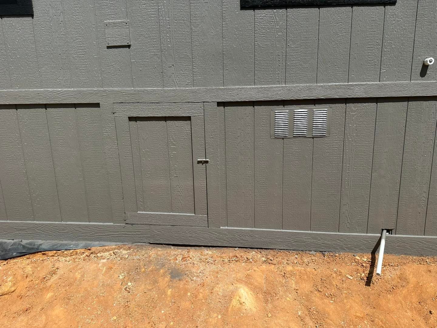 Side of a gray building with vertical siding and trim, set on a dirt foundation.