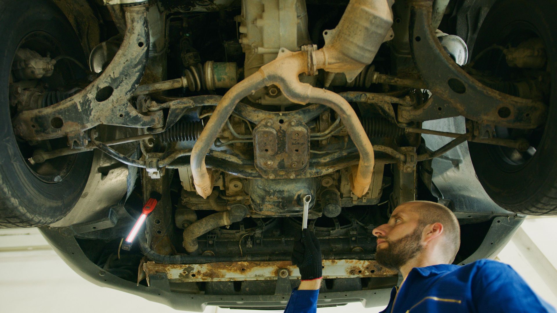 Mechanic inspects undercarriage of a car with a wrench and flashlight.