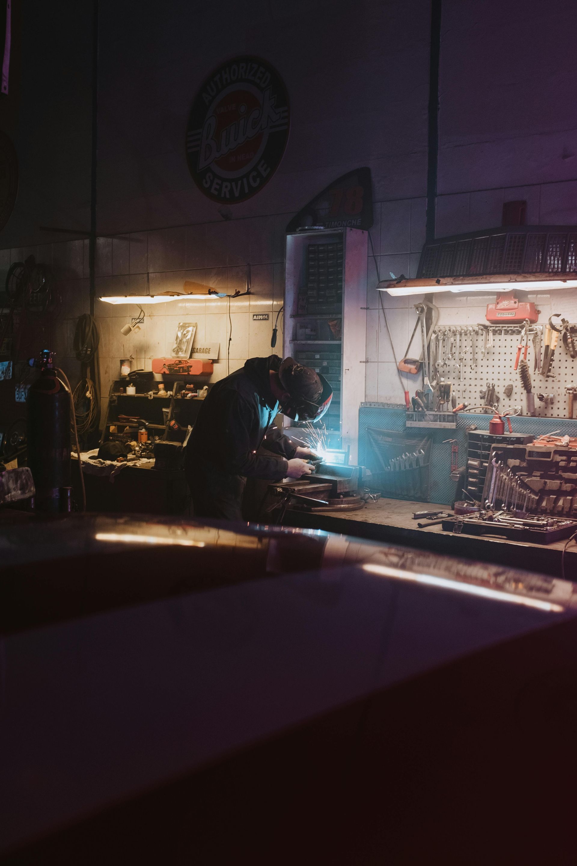 A person welding in a workshop, sparks flying, lit by neon and work lights.
