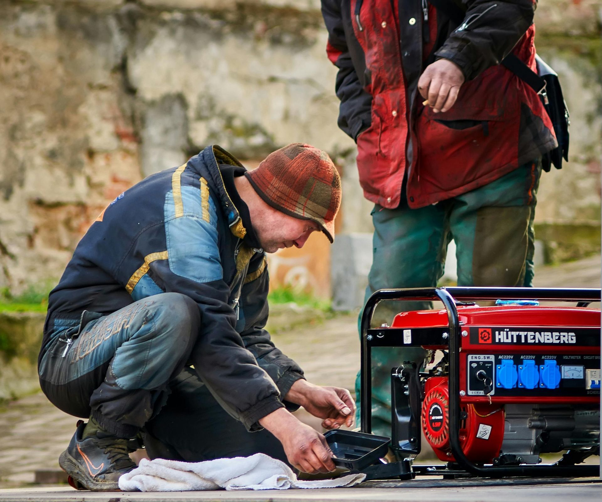 Man crouching, tending to red generator; another person looks on outdoors.