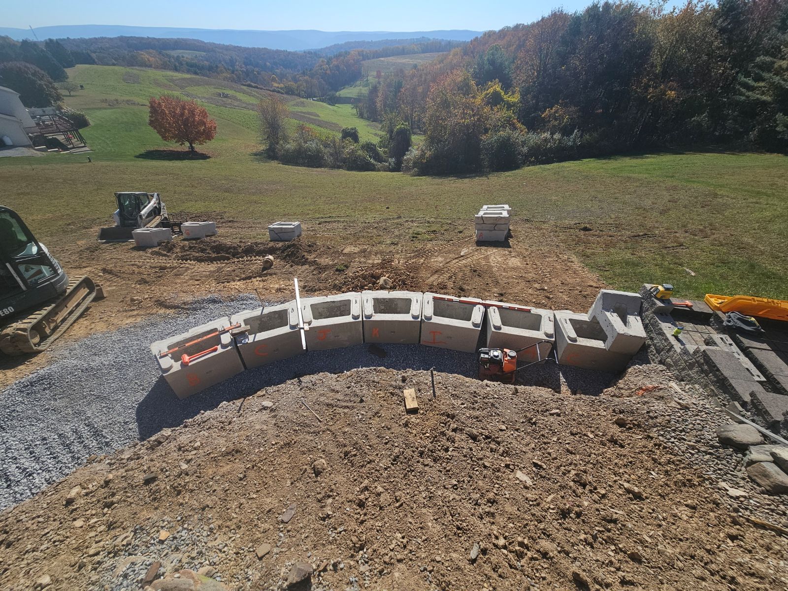 Curved cinder block wall under construction on a gravel lot overlooking a grassy valley and trees