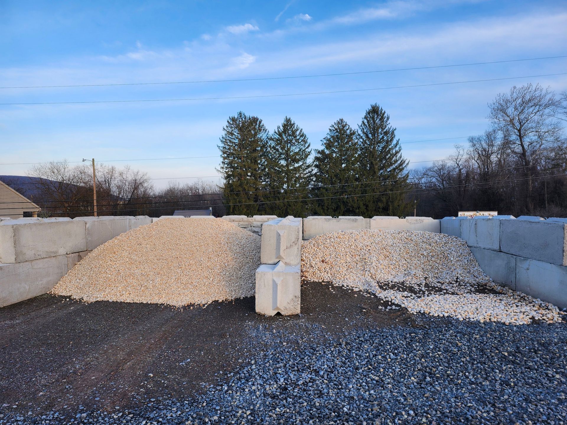 Piles of white gravel in a stone yard, with trees and a blue sky in the background.