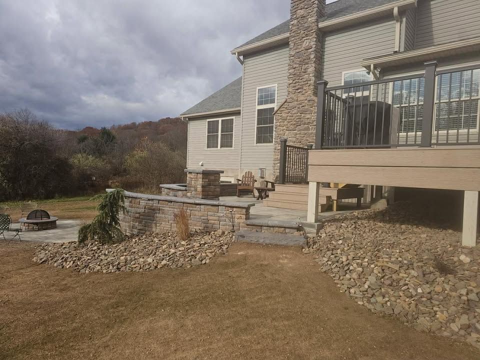 Stone patio and raised deck beside a beige house, with steps, retaining wall, and cloudy sky.