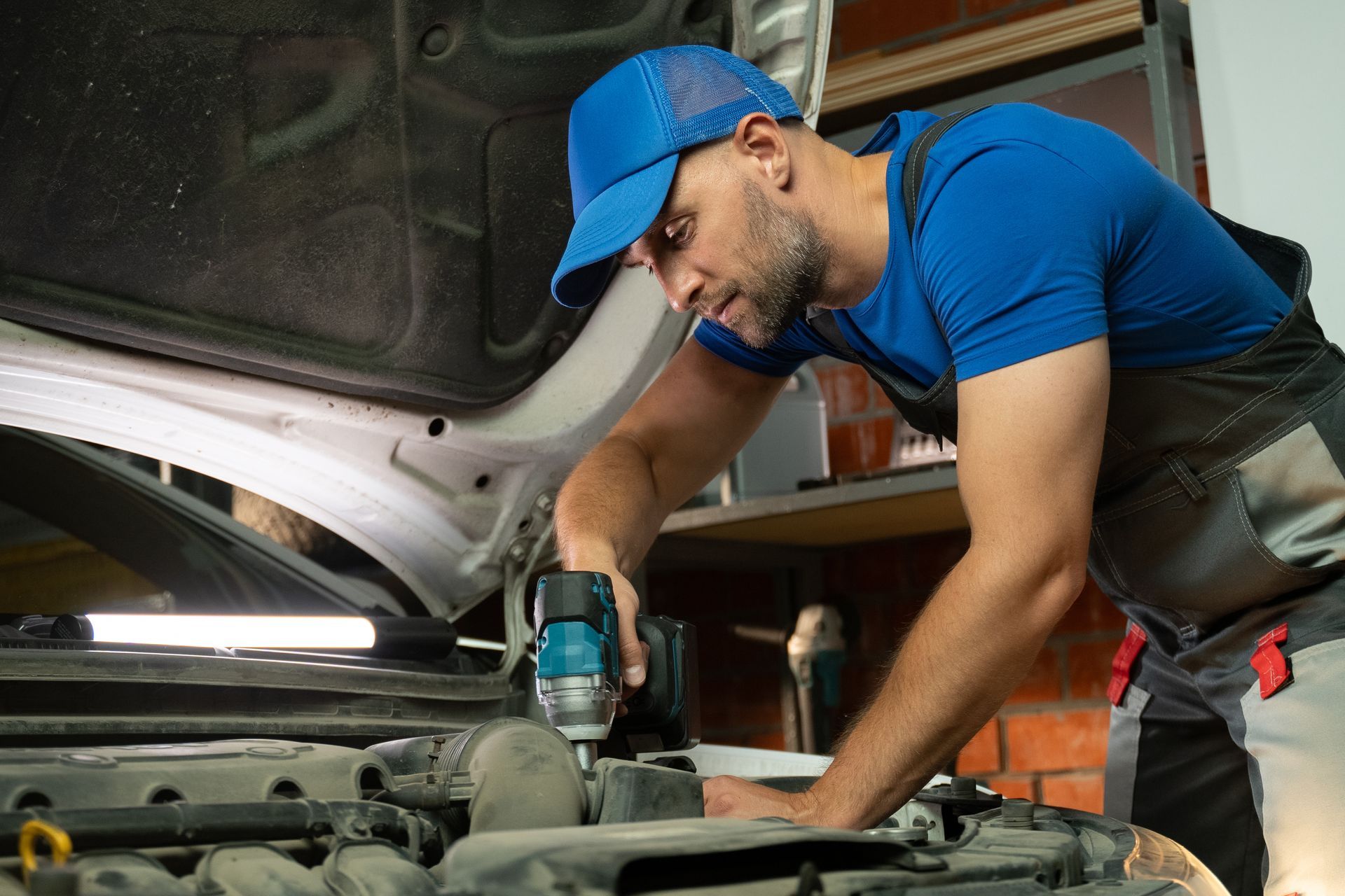 A mechanic works on a car engine in a garage. He is using a power tool to tighten bolts and screws.