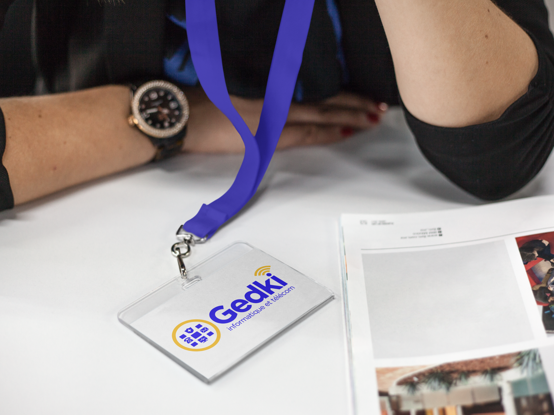 Une personne portant une montre avec un cordon retenant un badge Gedki, assise au-dessus d'un bureau et d'un magazine ouvert.