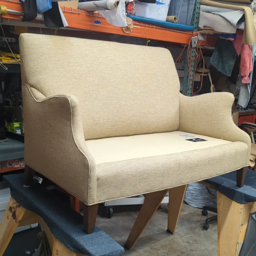 Beige upholstered armchair with curved arms in a workshop, resting on a wooden frame.