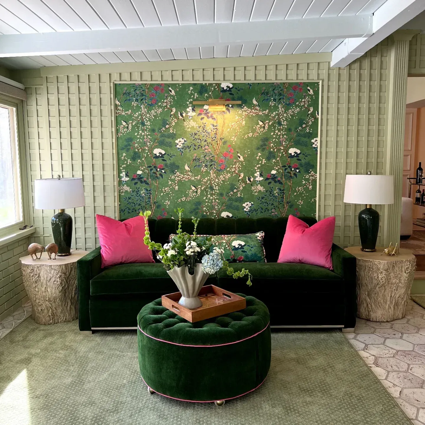 Green velvet sofa with pink pillows in a bright room, flanked by lamps and a round ottoman, with floral wall art.
