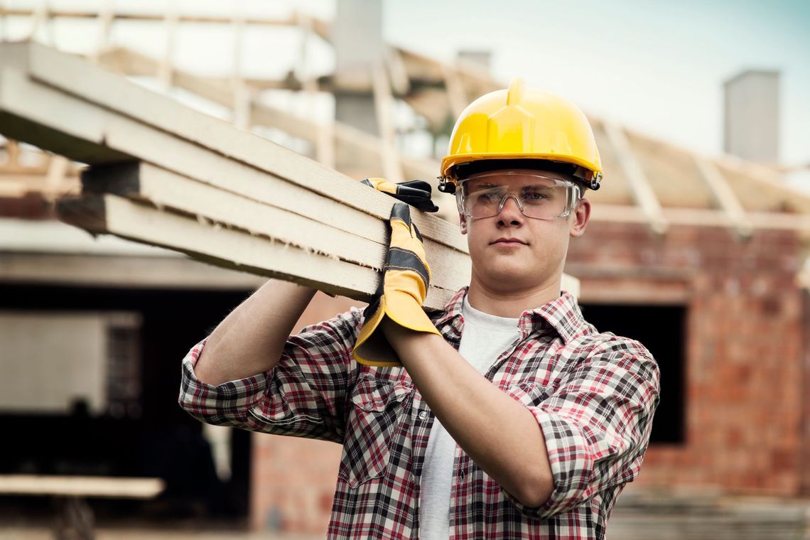 A construction worker is carrying a wooden beam on his shoulders.