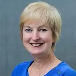 Blonde woman smiles, wearing a blue shirt, set against a grey-blue background.