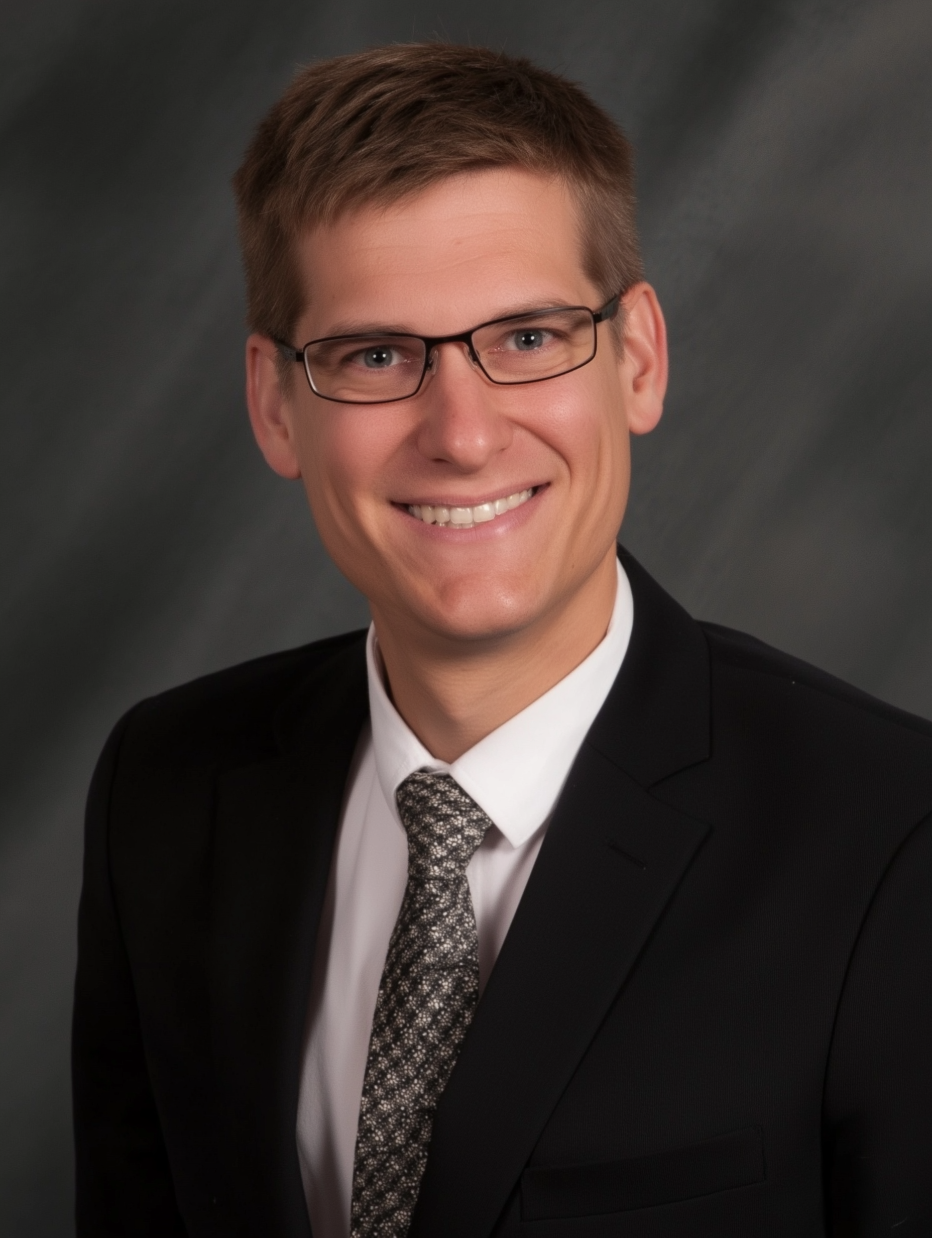 Man in a suit smiles, wearing glasses, dark background.