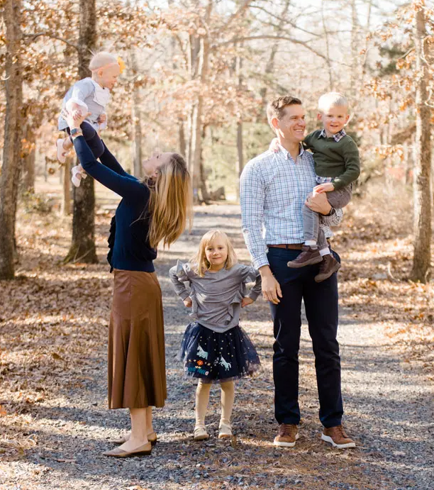 Family of five poses outdoors; parents holding children, one child standing. Autumn colors.