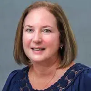 Woman with shoulder-length brown hair smiling in front of a gray background, wearing a blue top with lace detailing.