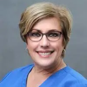 Woman with glasses and short blonde hair smiles in a blue shirt.