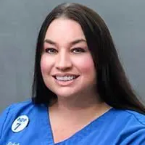 Woman with dark hair, wearing a blue scrub top, smiling.