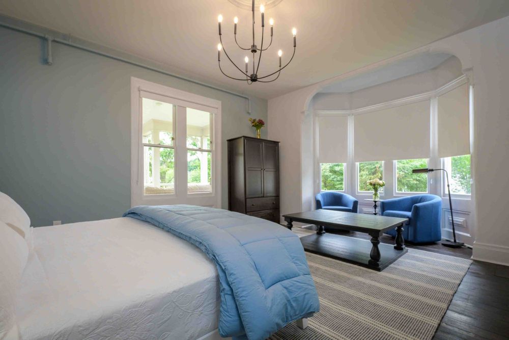 Bedroom with bed, blue blanket, armchairs, and bay window.