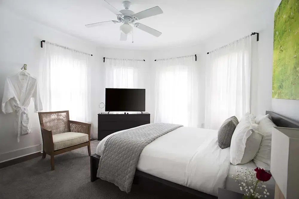 Bedroom with white walls, bed, chair, TV, and sheer curtains. A robe hangs on the wall.