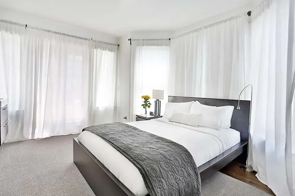 Bedroom with white curtains, gray bed, and a small table with a lamp and flowers.