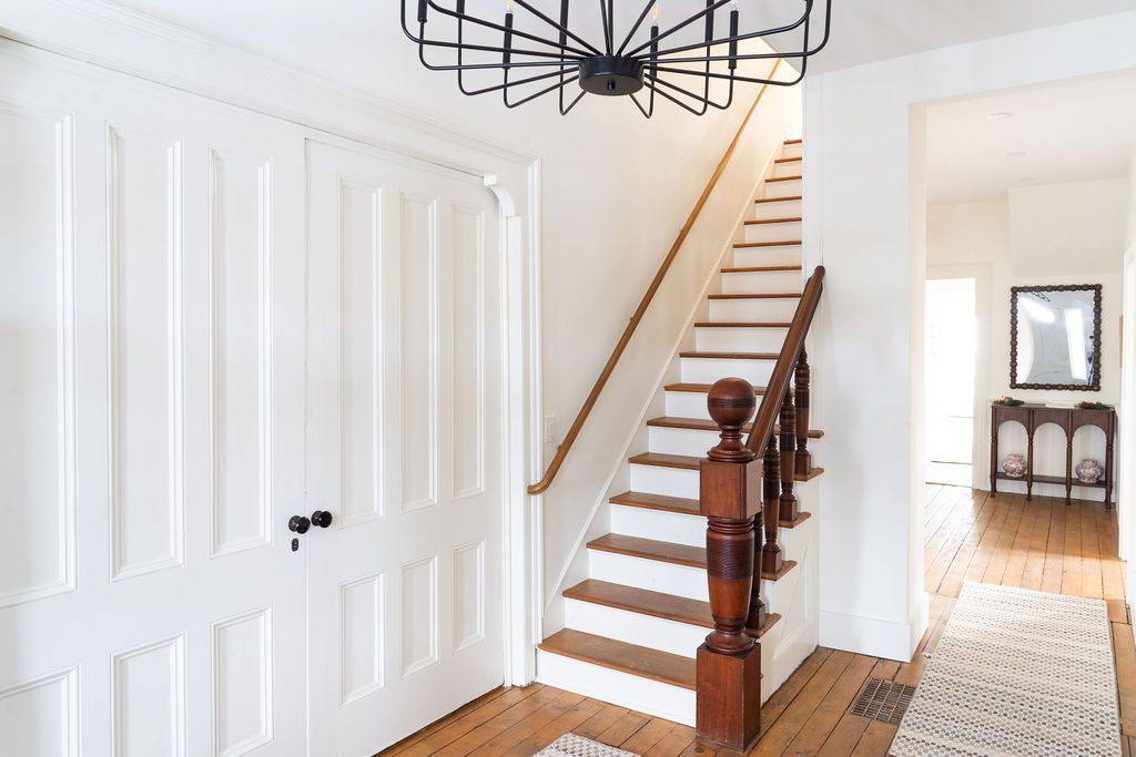 White hallway with staircase, wood floors, and modern chandelier.