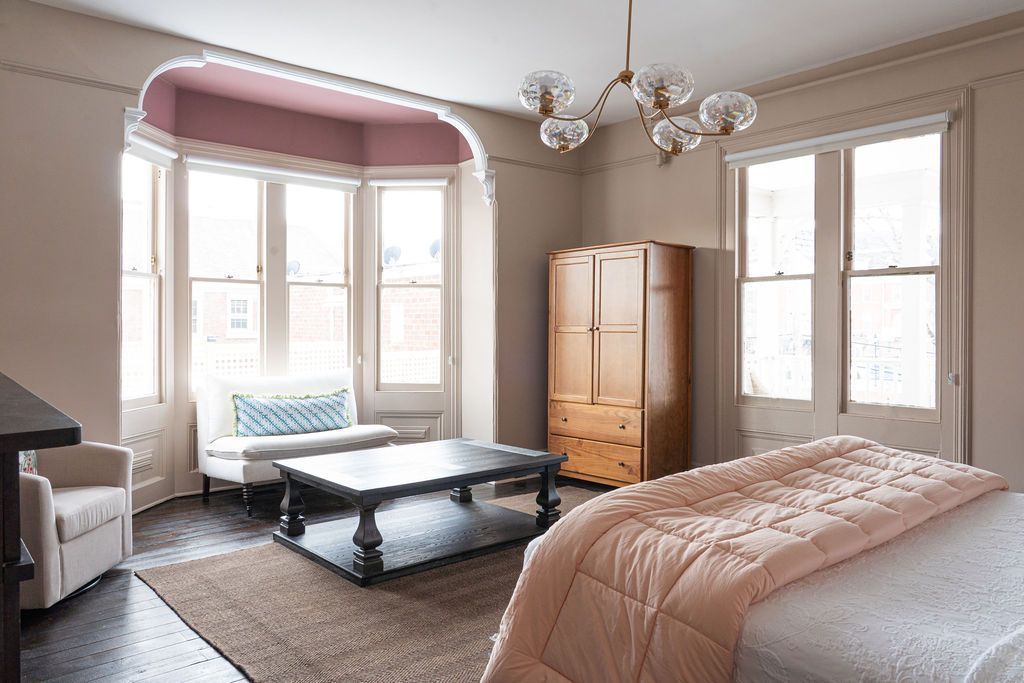 Bedroom with bed, seating area, and wooden wardrobe. Pale pink and neutral tones.