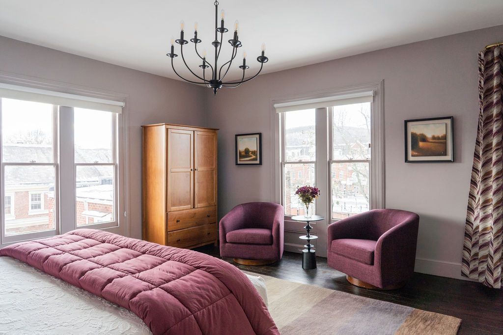 Bedroom with bed, armchairs, wardrobe, and chandelier. Walls are lavender, with dark wood floors.