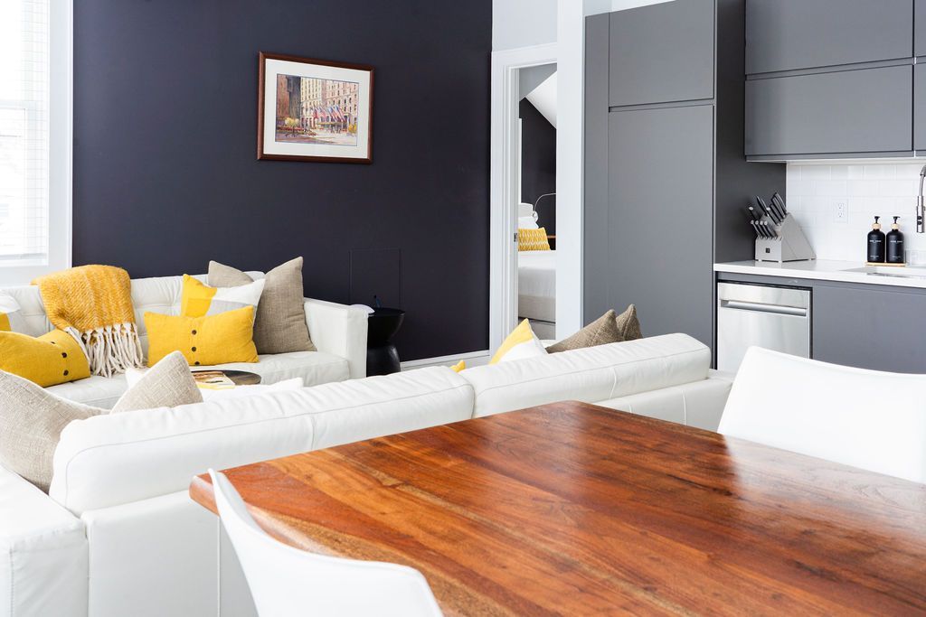 Modern living room with white sofas, dining table, dark accent wall, and multiple windows.