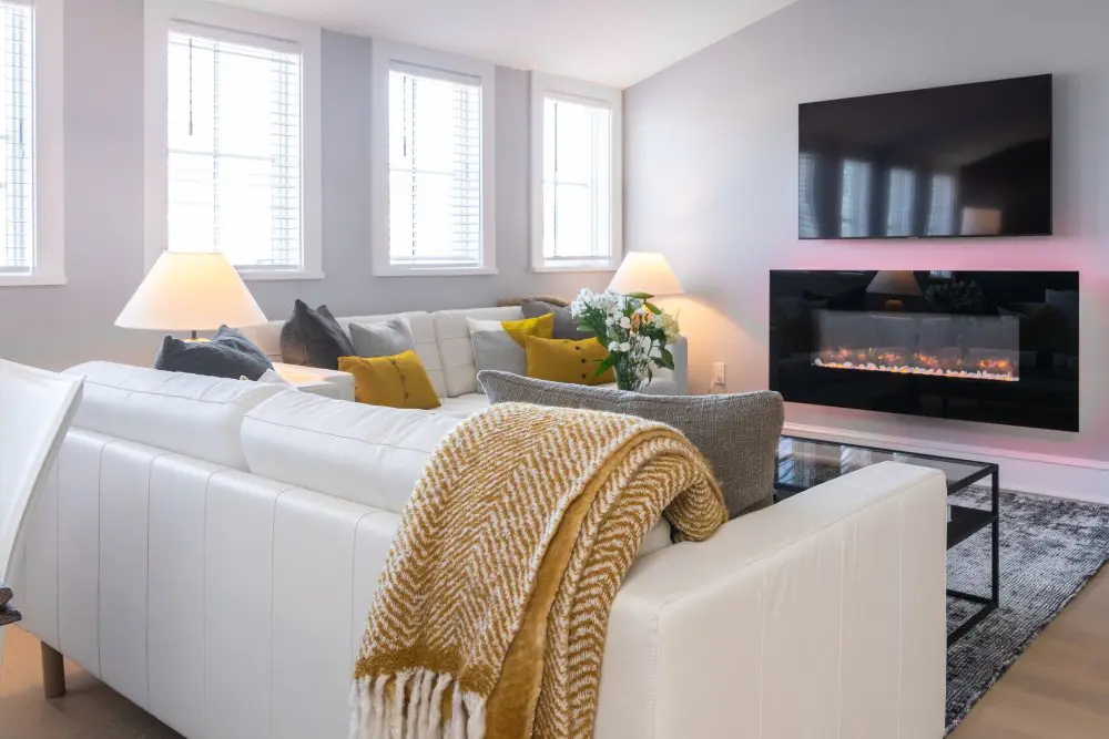 Cozy living room with white couch, fireplace, TV, and sunlight streaming through windows.