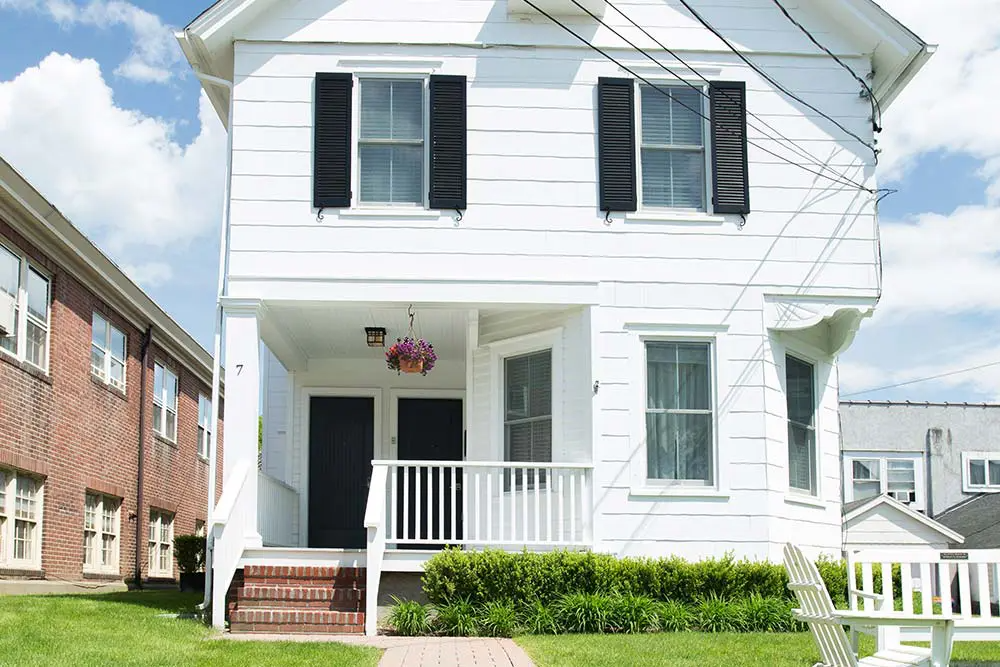 White two-story house with black shutters and a small porch. Red brick pathway and a green lawn.