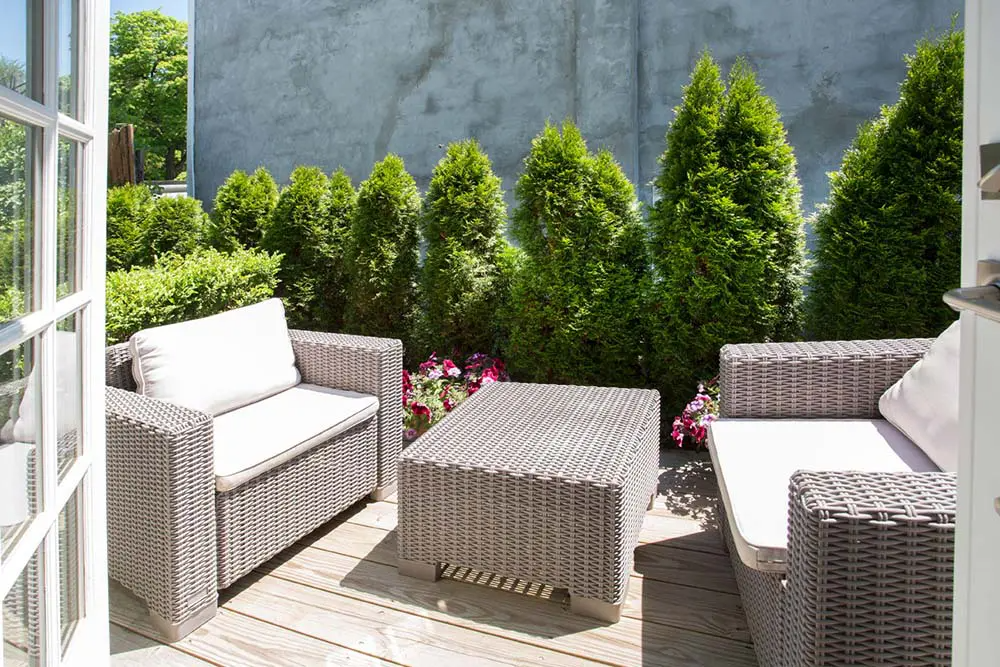 Wicker patio furniture on wooden deck, backed by green trees and grey wall. Open white door frame on left.