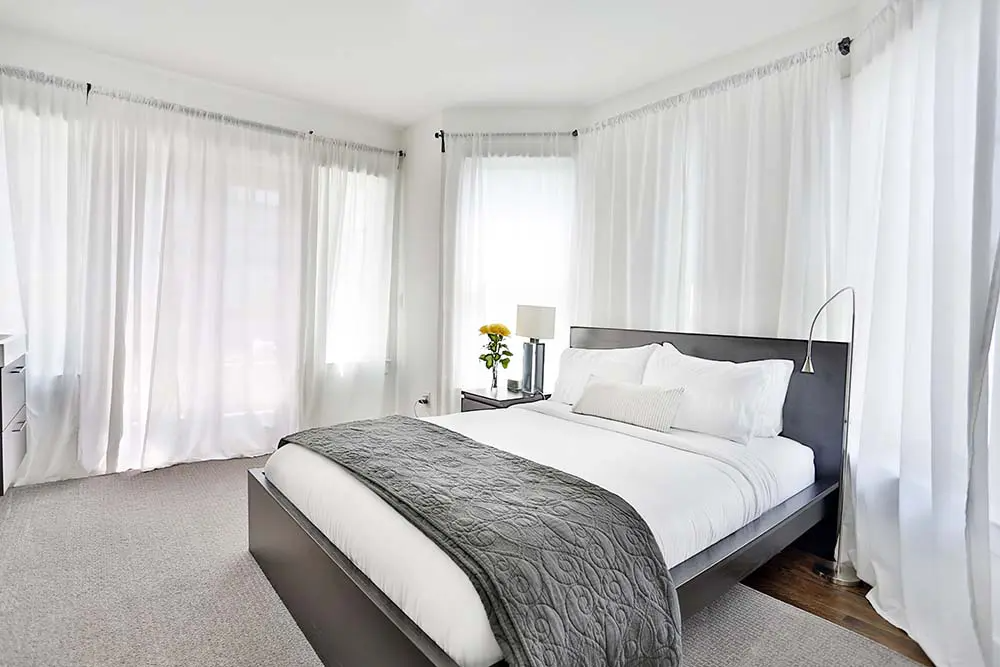 Bedroom with white curtains, gray bed, and soft rug.