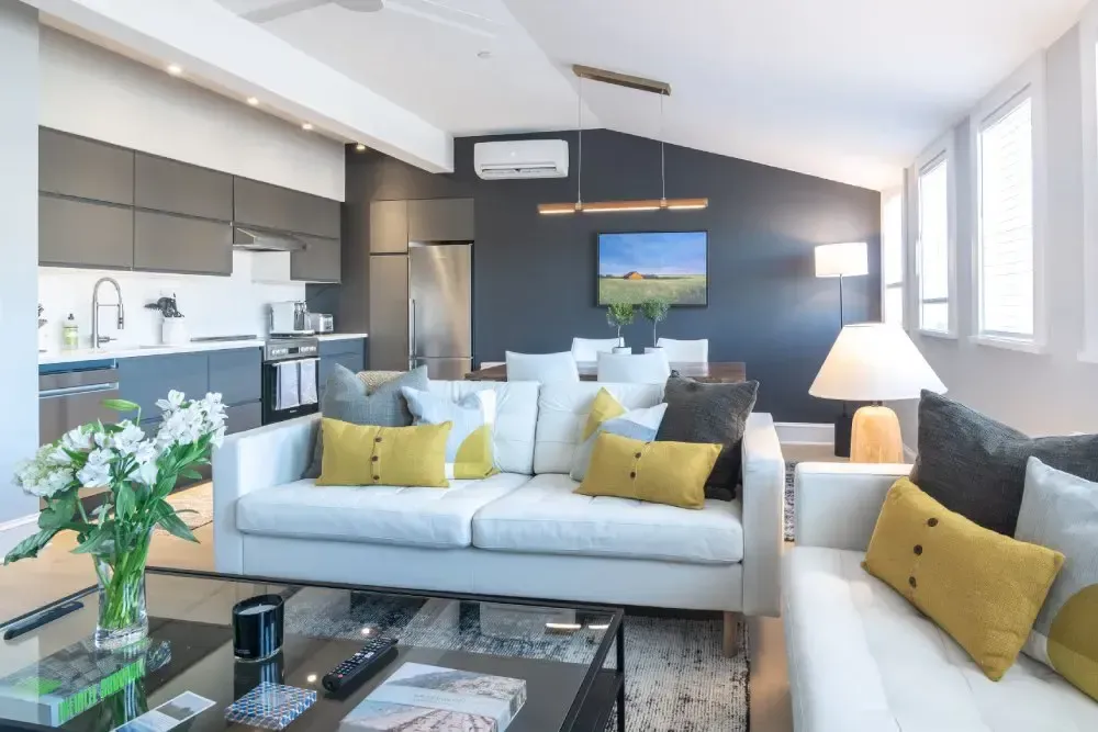 Open-plan living space with kitchen and seating area. White sofas with yellow pillows, dark gray kitchen cabinets