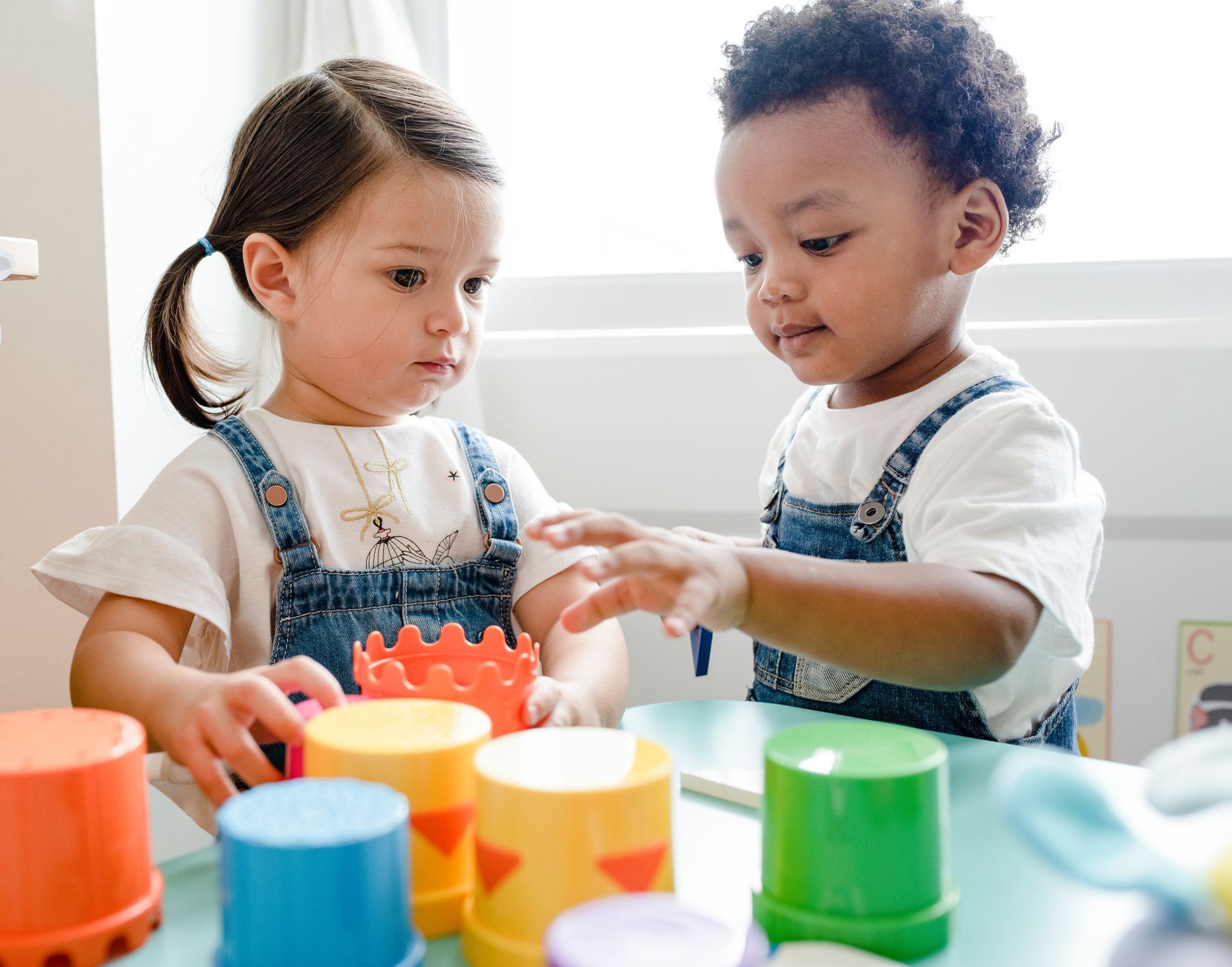 Two children playing with colorful stacking cups at a table, one reaching toward the toys