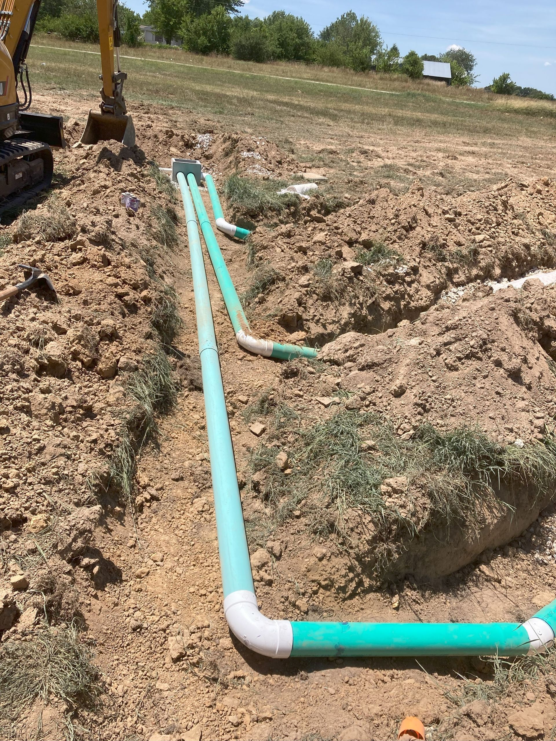 A green pipe is laying in the dirt next to a yellow excavator.